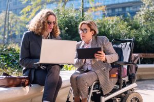 Two lawyers, one using a motorized wheelchair, review legal documents on their laptops outside