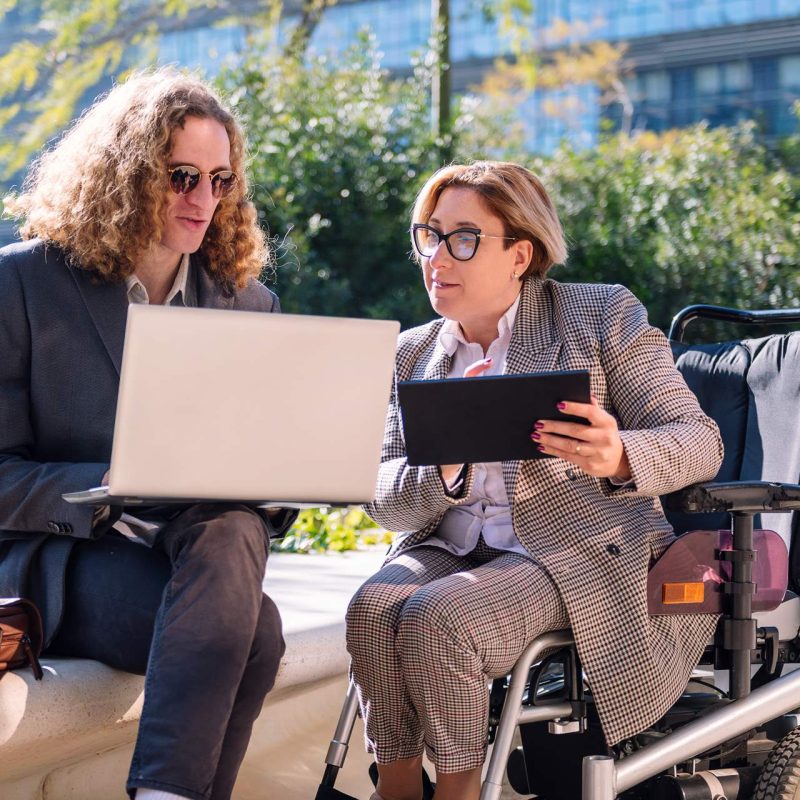 Two lawyers, one using a motorized wheelchair, review legal documents on their laptops outside