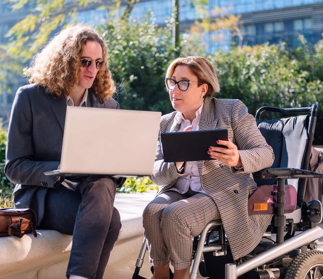 Two lawyers, one using a motorized wheelchair, review legal documents on their laptops outside